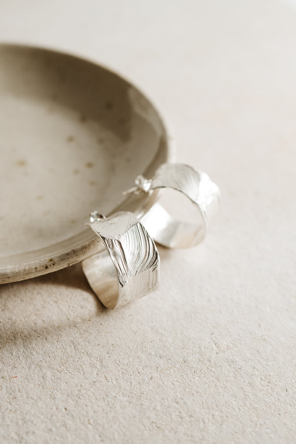 A pair of silver hoop earrings resting on a beige surface, with a focus on the textured surface of the hoops.