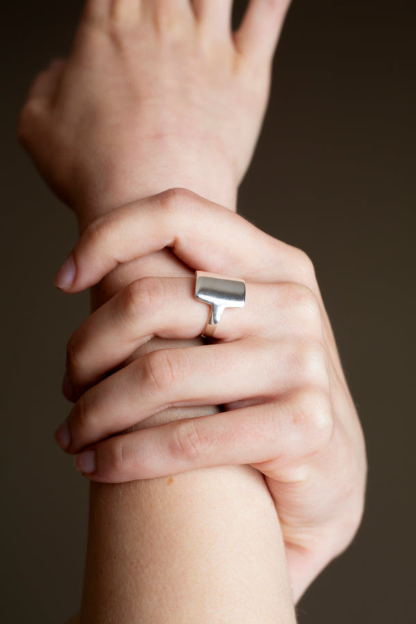 A person wearing a silver square shield ring on their finger, with the ring's bold, simple design visible against their skin.