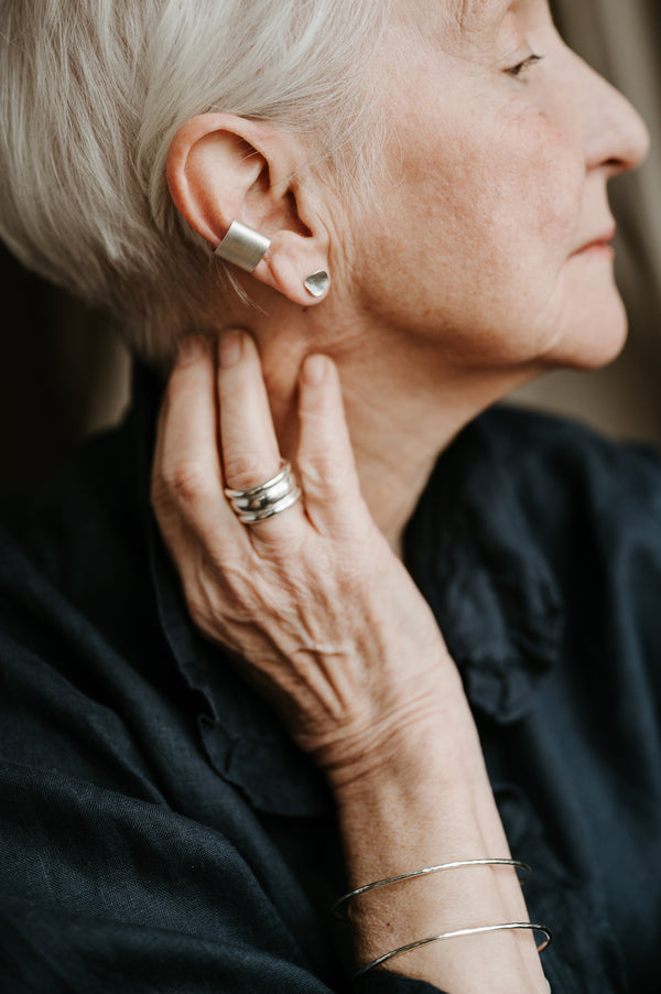 An older woman with short silver hair is featured, wearing the ear wrap which is silver and sits around the mid ear cartilage.