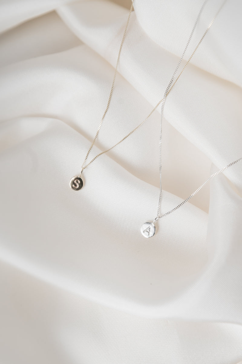 Two silver necklaces with small round pendants on a soft white fabric background