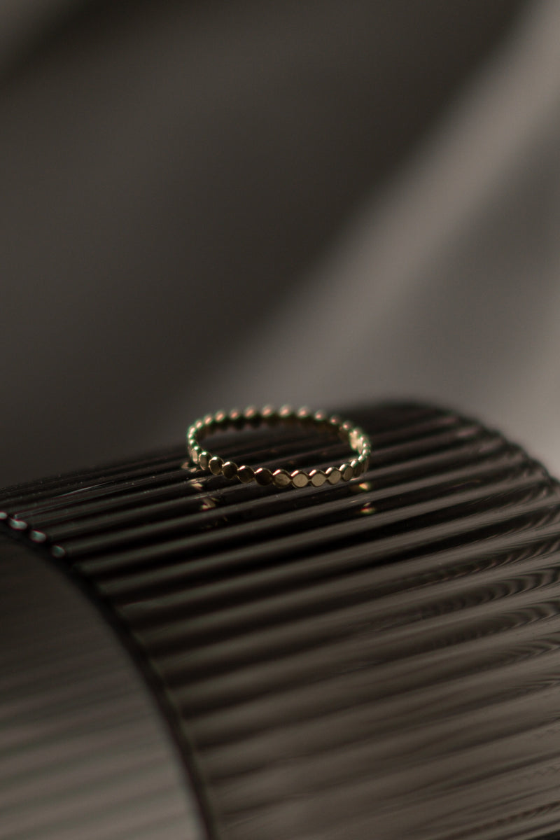 A close-up image of a gold stacking ring with beaded detailing, placed on a textured surface.