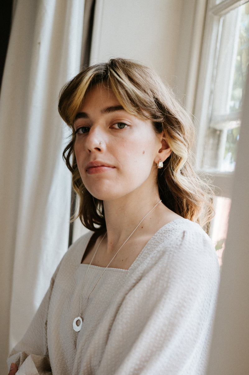 Woman wearing a light gray sweater standing indoors with a window in the background