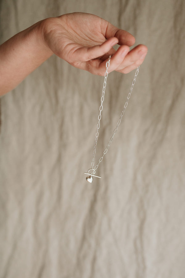 A person holding a silver necklace with a solid heart charm attached to a feature link and bar closure mechanism.