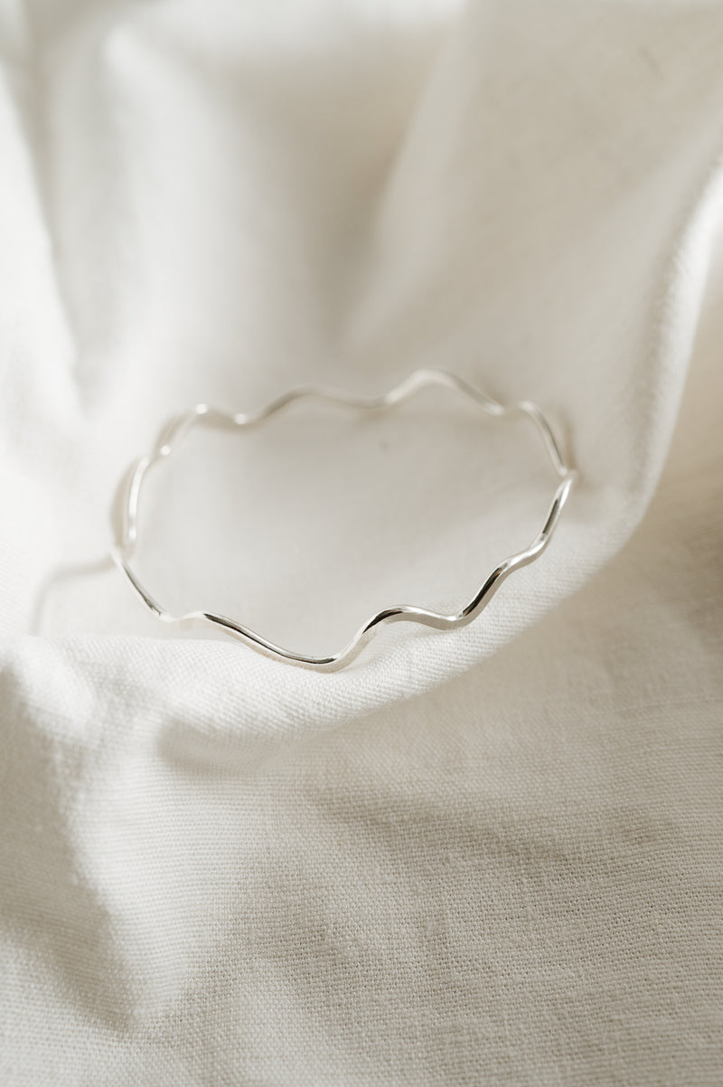 A silver bangle bracelet with a wavy design, displayed on a white fabric surface.