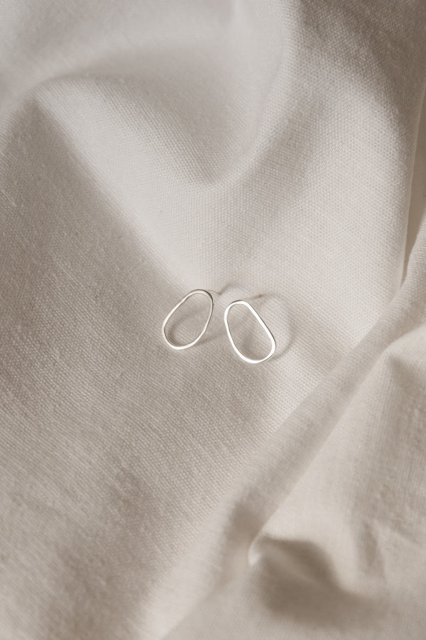 A pair of minimal organically shaped mini stud earrings displayed on a white fabric surface.
