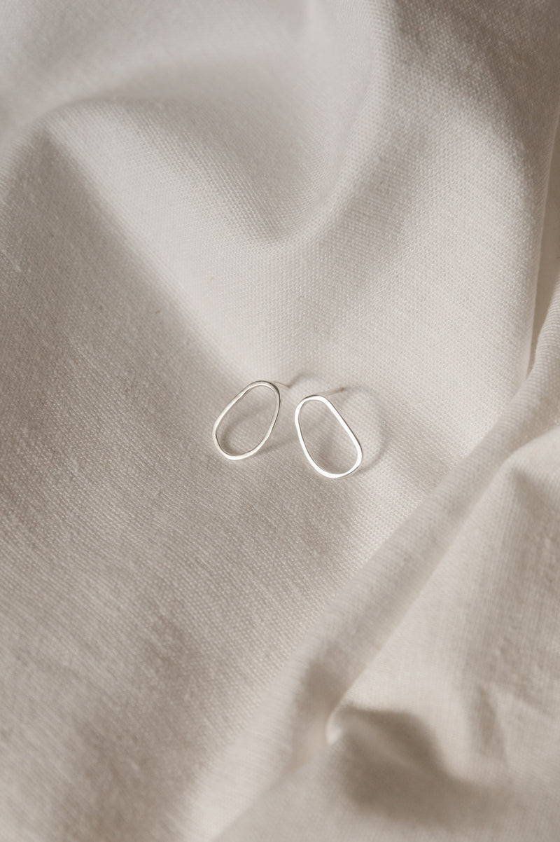 A pair of minimal organically shaped mini stud earrings displayed on a white fabric surface.