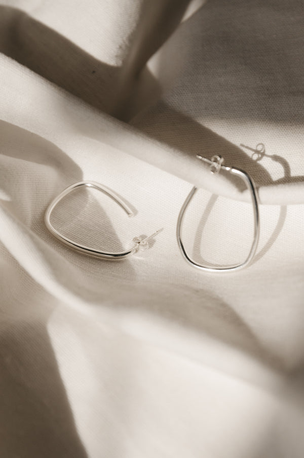 A pair of silver squoval hoop earrings with a satin finish, displayed on a white fabric surface.