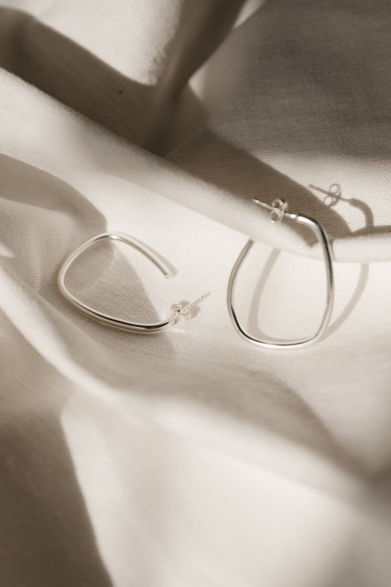A pair of silver squoval hoop earrings with a satin finish, displayed on a white fabric surface.